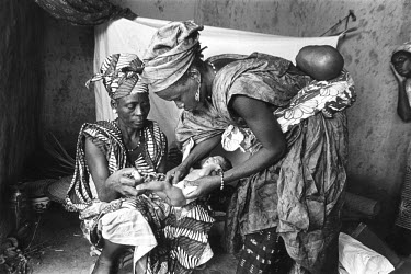 Village midwife with her own baby on her back and her assistant (left) inspecting and caring for a baby, only a few days old. In the right hand corner is the mother.