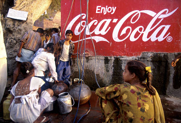 Adults and children collecting water from a communal water tap, next to an advertising billboard for Coca Cola.