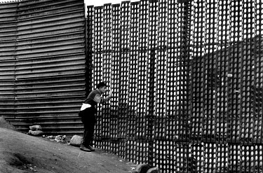 A migrant peers through the fence which marks the American-Mexican border, waiting for an opportunity to cross unheeded.