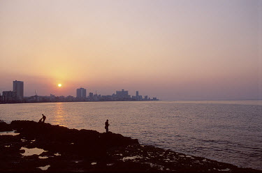 Men fishing from rocks as the sun sets on the city skyline.