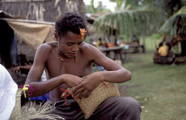 Youth with straw bags of cigarettes and betel nuts in his bachelor house. Kiriwina.