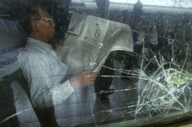 Businessman reading newspaper in Executive Class carriage of express train, next to window smashed by stone thrown by discontented peasants.