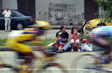 Competitors in the Vuelta a Colombia flash past a spectating family.  Political graffiti on the wall behind. The 53rd Vuelta a Colombia took place against a backdrop of civil war and violence. The bic...