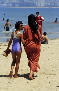 A mother - in traditional veil - and daughter, wearing a swimming costume, on the beach.