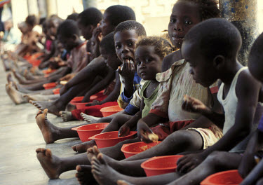 Children in a camp for internally displaced people (IDPs) eat a midday meal, prepared with food supplied by foreign aid agencies. An old gutted building provides shelter and schooling to people who ha...