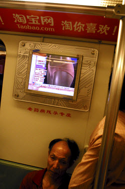 A man on a subway train sits beneath a video advertisement.
