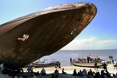 A rusting ship, with a small election poster, in the port of Beira. Fishermen rest in the shadows and launch small fishing boats from the beach.