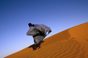 A man wearing a boubou, a traditional Hassaniya outfit, walks over a desert sand dune overlooking Chinguetti.