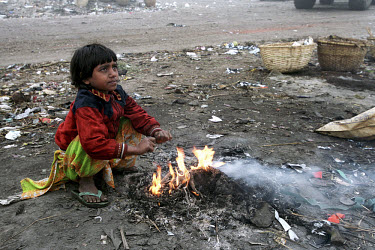 7 year old Jasmine warms herself beside a fire in the early morning on the Kajla rubbish dump where she works collecting saleable refuse.  The Kajla rubbish tip is one of three landfill sites in this...