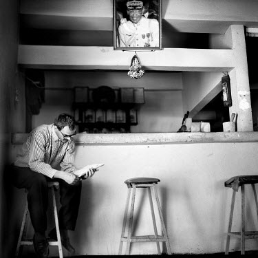 Young man reading a book on a stool in a hotel.  A photograph of King Hassan II hangs above the reception desk.