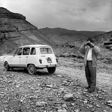 A man standing next to his car takes a photograph of the landscape.