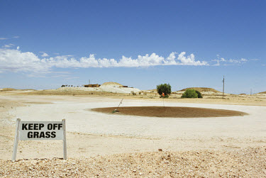 A sign reading 'Keep off the grass' stands at the edge of a parched putting green on a dry golf course.