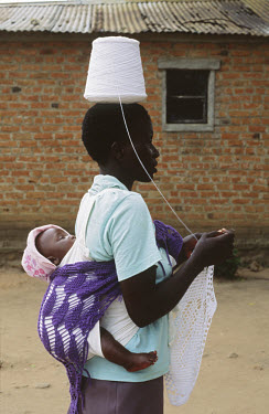 A woman knitting while carrying her baby on her back and a spool of wool on her head.