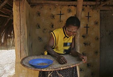 Religious and decorative symbols adorn the walls behind a young woman who is cleaning millet for the evening meal in a small roadside village.