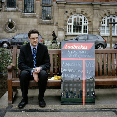 A man sits on a bench in Abingdon Street Gardens beside a board displaying bookmaker Ladbrokes' betting odds for the 2005 General Election. Abingdon Street Gardens, often referred to as College Green,...