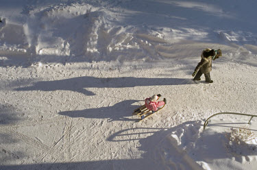 After the short days of the winter months, the March sun begins to throw long shadows on the snow as a mother pulls her daughter on a sledge to kindergarten.