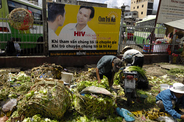 People picking over scraps of food from a market by an AIDS poster concerned with combatting discrimination at the main bus station in Cholon.
