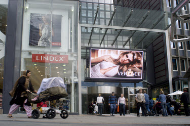 An immigrant mother with her child hurries under a billboard picturing a glamorous Versace model.