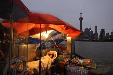 A sleeping sentinel at his stall on the Bund, decorated with an awning advertising Coca-Cola, as dawn breaks with Pudong financial district across the Huangpu Rier behind.