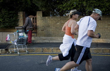 Morning joggers pass Joseph Maarman as he scratches through rubbish bins in a Green Point neighbourhood. Maarman, who lives in the bushes next to the Green Point Stadium, sells items he finds to raise...