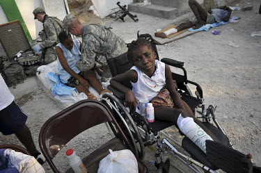 An injured girl sits in a wheelchair outside among the many others receiving treatment at the University Hospital five days after the earthquake hit Port-au-Prince. Behind her, US Army soldiers treat...
