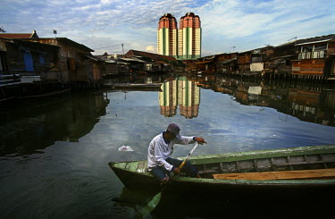 Man in a wooden boat navigating a polluted waterway backed by wooden slum housing, in contrast to the high rise appartment buildings visible behind.