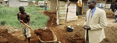 A businessman supervises a man working in a slum area building a new road in Kampala.