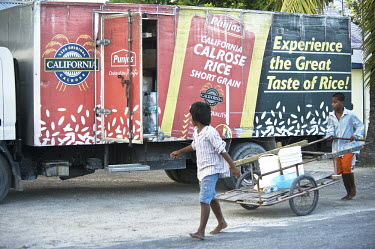 Boys wheel a cart carrying containers that they will fill with fresh water and walk past a truck with signage advertising imported rice.