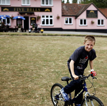 A boy rides his BMX bike across a grass field in front of the Five Bells Pub, a country pub in Cavendish, Suffolk.