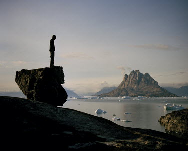 A child stands on a rock looking out over floating pieces of ice towards the Uummannaq Mountain.