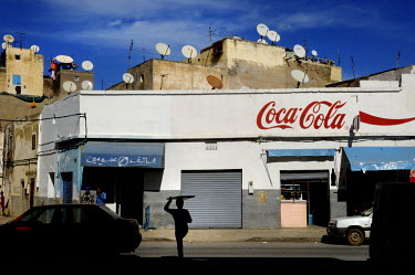 A boy delivering tea is silhouetted against a shop front that is adorned with a hand painted Coca Cola advertisement. The roofs above the shop are lined with satellite dishes.