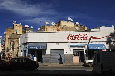 A man in a burnoose walks past a Coca Cola advertisement painted on a shop front. The roofs above the shop are lined with satellite dishes.