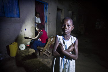Christine Akurut's daughter stands with her arms crossed, while her mother, a primary school teacher, prepares a meal under the light of the Firefly, a portable LED lamp powered by solar energy.