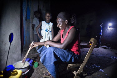 Christine Akurut's daughter watches on while her mother, a primary school teacher, prepares a meal under the light of the Firefly, a portable LED lamp powered by solar energy.