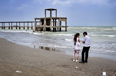 A couple argue while standing on beach by the Caspian Sea. In the background is the skeletal remains of a Soviet era restaurant.