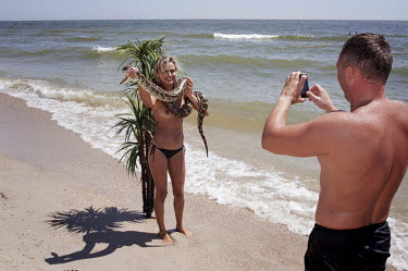 A female tourist, holding a rented snake, poses for a photograph on a beach in front of a plastic palm tree in the cheap Black Sea resort destination of Zatoka.