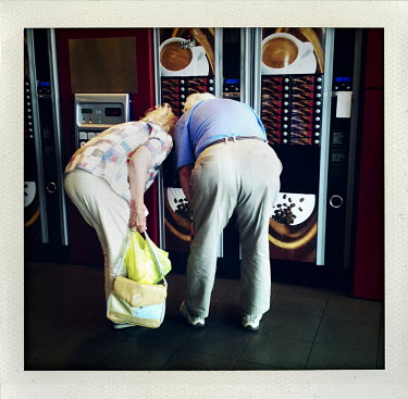 An elderly couple buy coffee from a vending machine.
