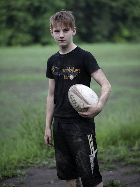 A young boy, covered with mud, stands in a field with a rugby ball.
