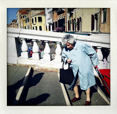 An elderly woman, pulling a shopping trolley, negotiates the steps of a canal bridge.