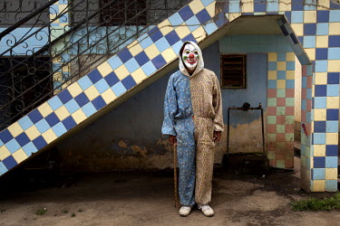 A clown stands outside a building, as people dance house to house, sharing food, during Carnival.