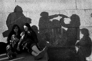 Two girls  sit on the embankment of the Neva river while their friends drink from a shared bottle.
