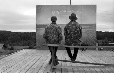 Two man sit on a bench in front of a banner which reads: 'We live on the Sea - Sing for the Sea!' in a village on the coast of the White Sea in the far North of Russia. This area is inhabited by the P...