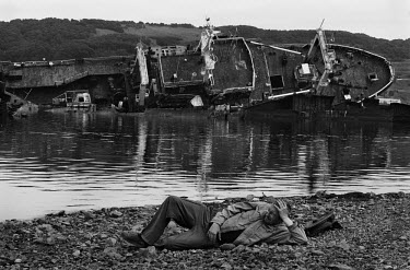 A man lies on a beach with a derelict boat on its side visible in the background by the sea in the Russian region of Kamchatka.