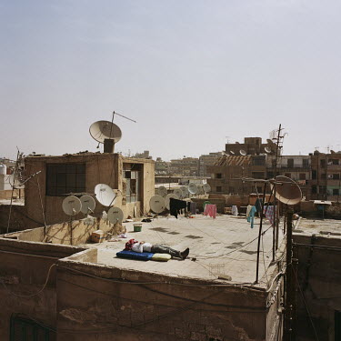 A man sleeps on a rooftop surrounded by television satellite dishes and drying clothes.