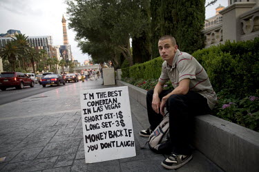 A young man sits with a billboard that advertises his skills as a comedian for hire on the 'Strip', in Las Vegas, Nevada.