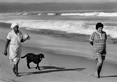 A black maid carries a dog's leash for her white employer on as they walk a dog on a whites only beach.