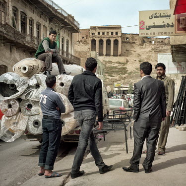 A young man sits on top of a pile of rolls of carpet loaded onto the back of a pick up truck below the ancient Citadel of Arbil while three other man stand nearby. The Citadel is an inhabited mound in...