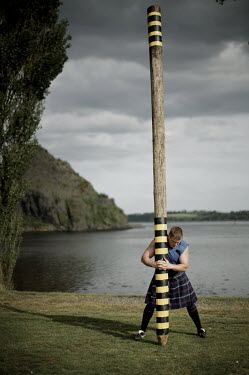 Neil Elliott, dressed in a kilt and holding a caber on a likeside near his home in Dumbarton. He competes in over 40 Highland Games and heavyweight events across the world throughout the summer.