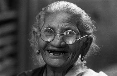 An elderly woman, wearing a pair of glasses with thick lenses, smiles a toothless smile.