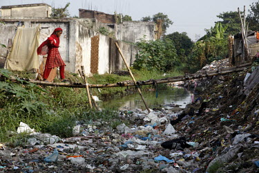 A young woman walks over a makeshift bridge that spans a rubbish strewn and polluted river which runs through the middle of a slum.   Walking across makeshift bridge which spans waterway/open sewer th...